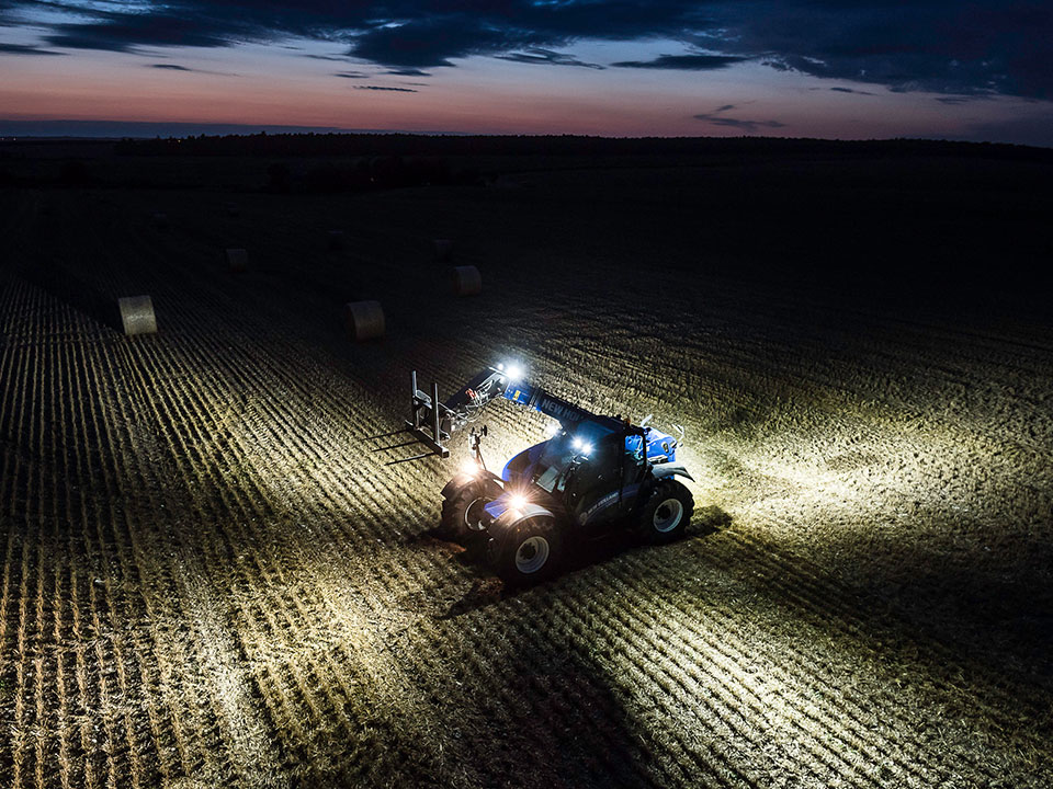 blauer new holland teleskoplader mit scheinwerfer bei nacht auf feld