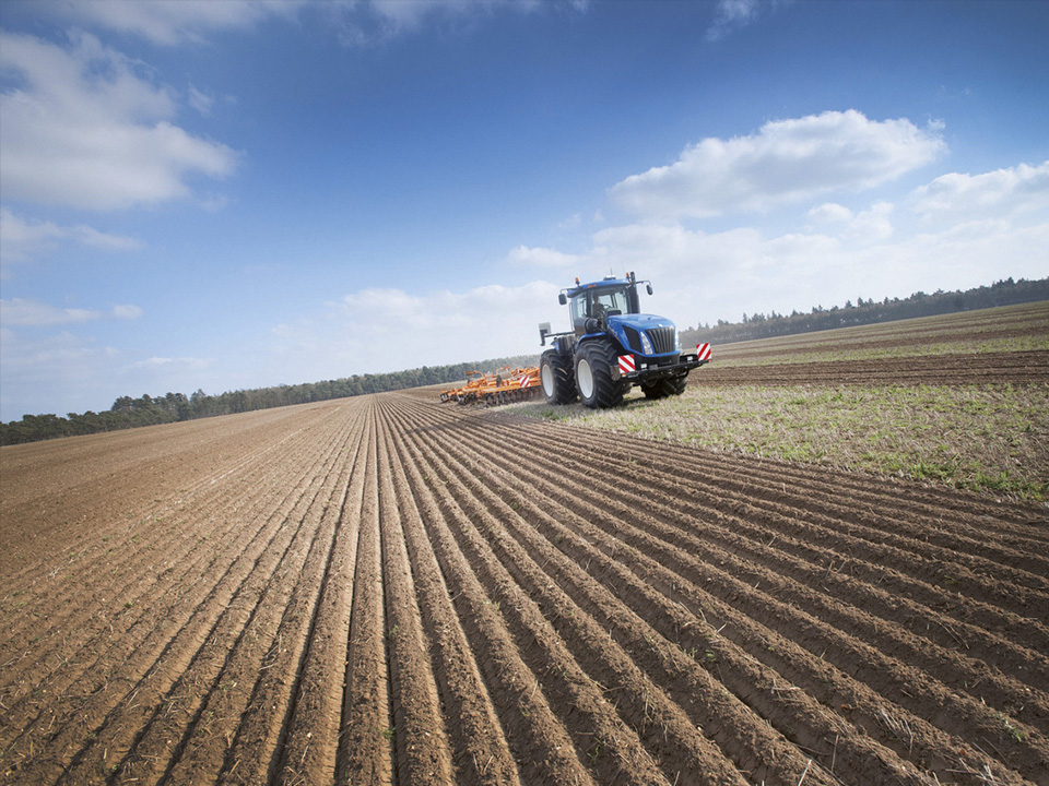 new holland traktor von vorne mit pflug auf feld blauer himmel mit wolken