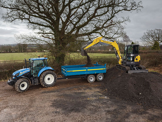 gelber new holland minibagger auf erdhaufen beläd blauen anhänger blauer new holland traktor