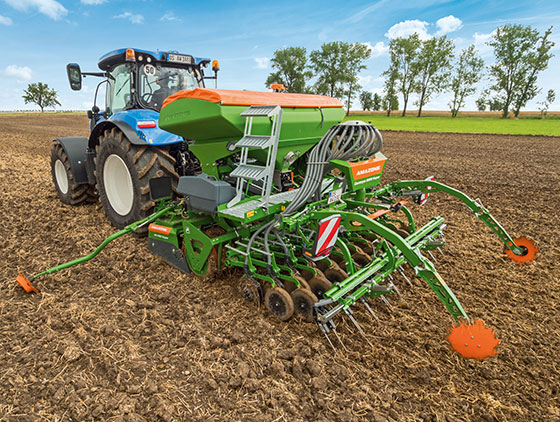 blauer traktor im seitlichen profil mit grün orangener amazone sämaschine auf feld bäume im hintergrund