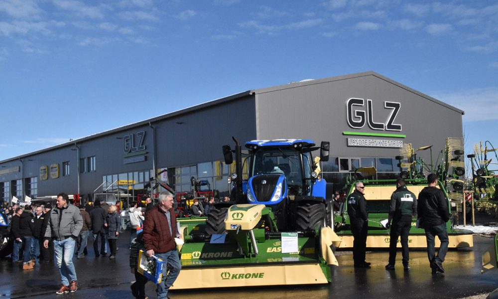 blauer new holland traktor mit grünem krone anbaugerät vor glz gebäude besucher schauen sich ausstellung an